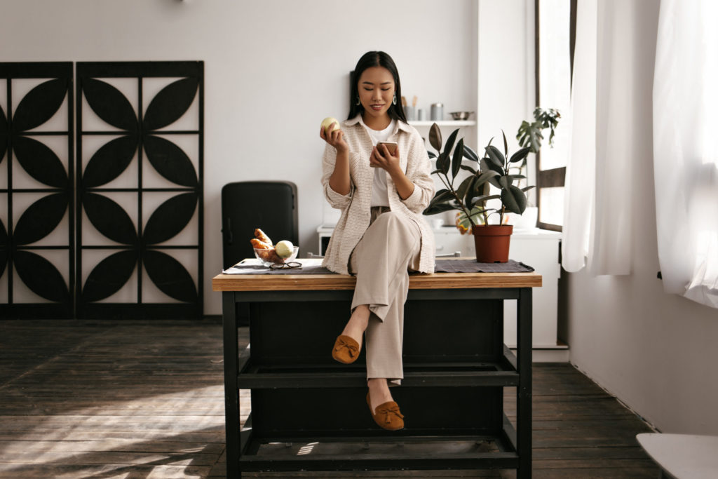 Happy attractive brunette woman in beige outfit sits on wooden table on kitchen. Asian lady in good